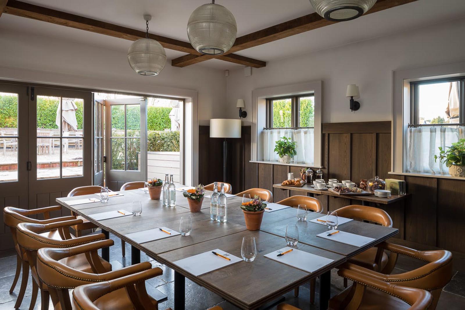 Hurley House - A cozy meeting room with a large wooden table surrounded by leather chairs. The table is set with placemats, glasses, and water bottles. Natural light streams through open double doors, and potted plants decorate the space.