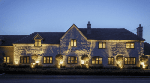 Hurley House - A cozy stone building, illuminated at dusk against a clear blue sky. Warm lights highlight the facade and garden, creating a welcoming atmosphere. The building features several windows and a gabled roof.