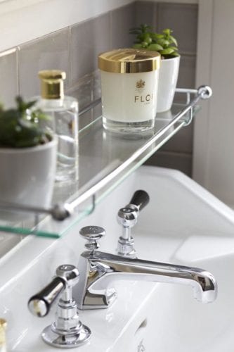 Hurley House - A bathroom sink with chrome faucets. Above it, a glass shelf holds a candle labeled FLO, a small potted plant, and a perfume bottle. The background features beige tiles.