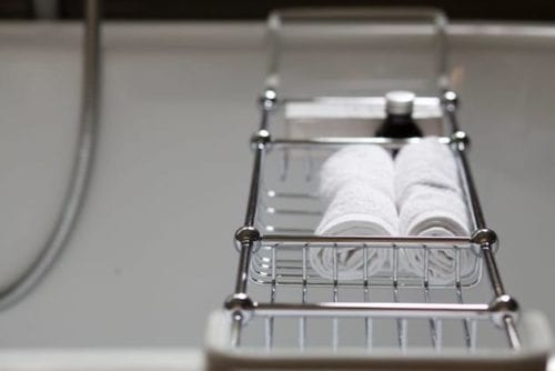 Hurley House - A chrome bathtub caddy holding two rolled white towels and a black bottle, placed over a white bathtub.