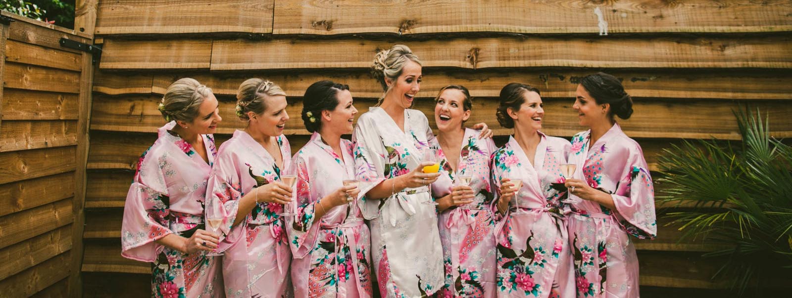 Hurley House - A group of seven bridesmaids in floral robes, six in pink and one in white, stand together laughing and holding drinks by a wooden fence. They appear to be celebrating at a luxury wedding venue in Berkshire, possibly during a pre-wedding event.