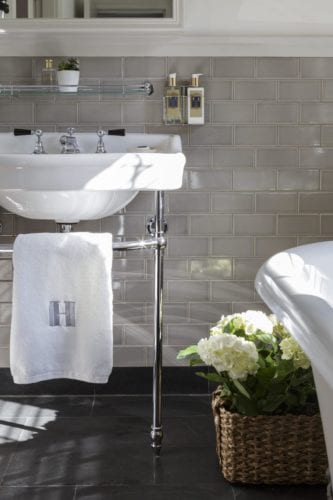 Hurley House - A bathroom with a white sink on chrome legs, set against a beige subway tile wall. A small plant is on a glass shelf above the sink, and a wicker basket with white hydrangeas sits on the floor. A white towel with an H monogram hangs from the sink.