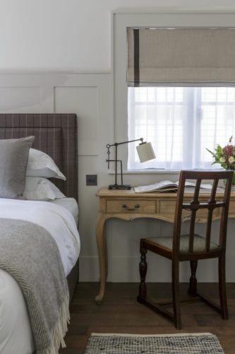 Hurley House - A cozy bedroom corner with a neatly made bed featuring a gray headboard and white linens. Adjacent is a wooden desk with an angled lamp and an open book, accompanied by a wooden chair. A window with sheer curtains lets in soft light.