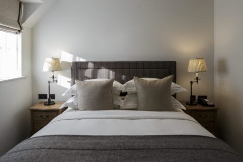 Hurley House - A cozy bedroom with a neatly made double bed, featuring a checkered headboard and two beige pillows. Two bedside tables hold matching lamps. Soft light comes through a window on the left, creating a warm atmosphere.