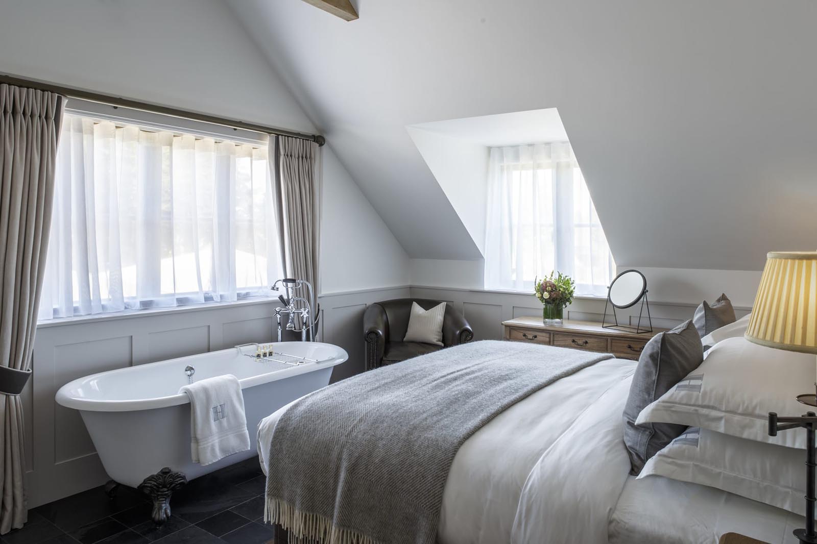Hurley House - A cozy bedroom with a clawfoot bathtub by the window, a large bed with gray bedding, a wooden dresser with a mirror and vase of flowers, soft lighting from a table lamp, and slanted ceilings with a skylight.