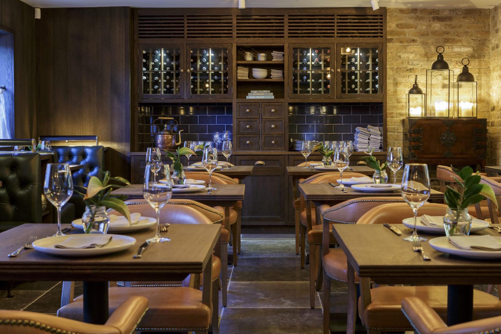 Hurley House - A cozy restaurant interior with wooden tables set for dining, featuring wine glasses, plates, and cutlery. Leather chairs complement the warm decor. In the background, a cabinet displays bottles and dishes, while soft lighting adds a welcoming ambiance.
