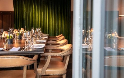 Hurley House - A well-set dining table in an elegant restaurant with wooden chairs and a green velvet curtain in the background. The table is adorned with wine glasses, white napkins, and small decorative flowers, creating a sophisticated atmosphere.