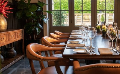 Hurley House - A warmly lit dining room with a long wooden table set for a meal. Leather chairs surround the table, which is adorned with wine glasses and cutlery. Large windows reveal greenery outside, and a wooden sideboard with a vase and plants is in the background.