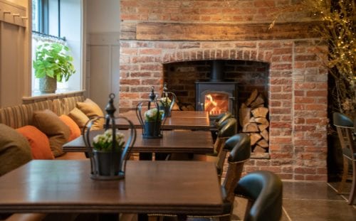 Hurley House - Cozy cafe with a rustic brick fireplace, a wood-burning stove, and stacked logs. Wooden tables with small potted plants and cushioned seating line the wall. Warm lighting adds to the inviting atmosphere.