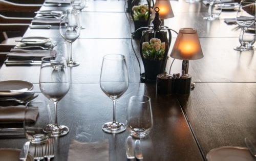 Hurley House - A long wooden table set for dining, with neatly arranged wine glasses, plates, and cutlery. Decorative lamps and small vases with flowers line the center, creating an elegant and cozy atmosphere.