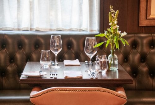 Hurley House - A neatly set dining table for two with wine glasses, water glasses, and cutlery on a dark wooden table. A small vase with white flowers decorates the table. The background features tufted leather seating and a curtain.