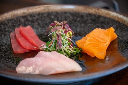 Hurley House - A black plate with three types of sashimi: slices of pink tuna, orange salmon, and pale white fish, garnished with microgreens in the center.