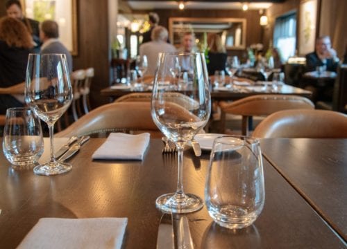 Hurley House - A dimly lit restaurant with elegantly set tables, featuring empty wine glasses and folded napkins. Patrons are seen in the background, creating a cozy and inviting ambiance.