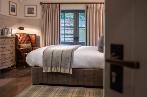 Hurley House - Cozy bedroom with a large bed, gray blanket, and plaid rug. A brown leather armchair and a wooden dresser are on the left. Soft light filters through curtains on a glass door, revealing a wooden fence outside. A lamp sits on the dresser.
