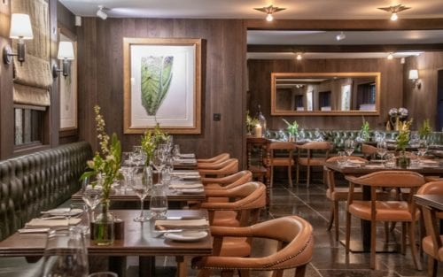 Hurley House - A cozy restaurant interior with wooden walls and floors, featuring leather chairs and tables set for dining. A large mirror and a framed leaf artwork adorn the walls, while small vases with flowers add to the ambiance.