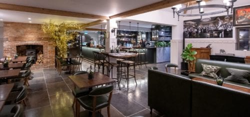 Hurley House - A cozy, modern bar and restaurant interior with wooden beams, a brick fireplace, and various seating arrangements. Green plants and framed photos add decoration. The bar area is well-lit and stocked with bottles.