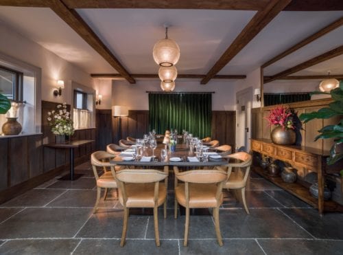 Hurley House - A dining room with a long table set for a meal. It features wooden beams, pendant lights, and a green velvet curtain. On the walls are mirrors and large vases with flowers. The wooden floor and warm lighting create a cozy atmosphere.