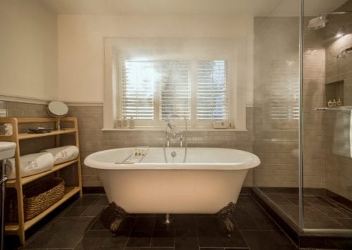 Hurley House - A cozy bathroom with a freestanding white bathtub on clawfoot legs, a wooden shelf with towels, a glass-enclosed shower, and a window with shutters. The room has beige tile walls and a dark tiled floor.