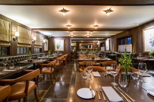 Hurley House - A cozy and elegantly furnished restaurant interior featuring wooden tables, leather chairs, and soft lighting. The tables are set with plates, cutlery, and glasses, surrounded by artwork and large windows with blinds.