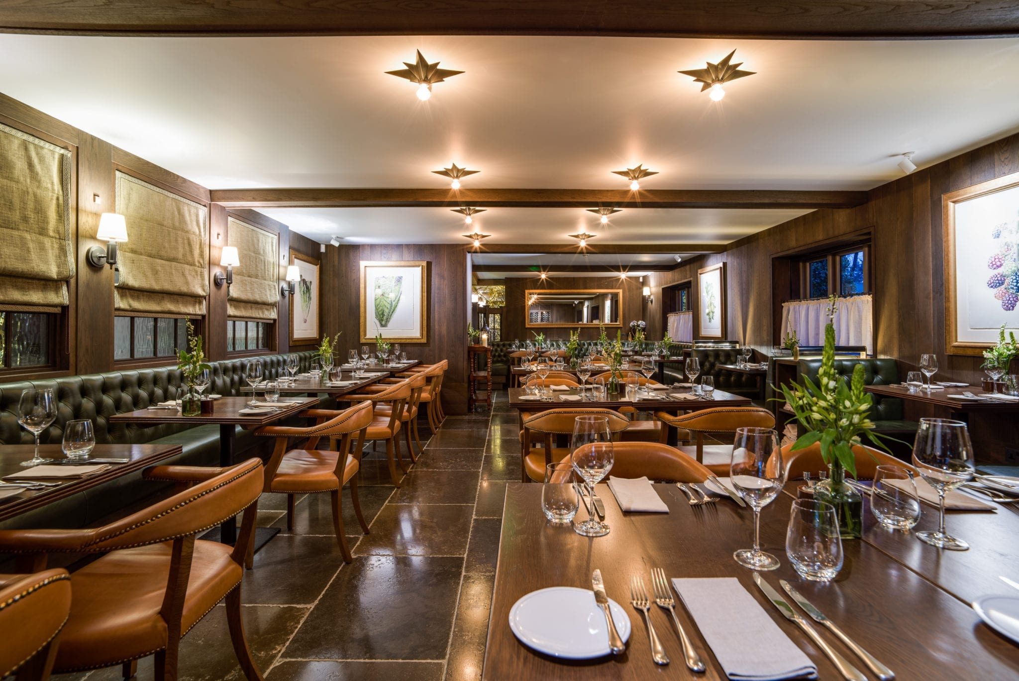 Hurley House - A cozy and elegantly furnished restaurant interior featuring wooden tables, leather chairs, and soft lighting. The tables are set with plates, cutlery, and glasses, surrounded by artwork and large windows with blinds.
