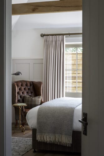 Hurley House - A cozy bedroom with a large window covered by beige curtains. A brown leather armchair sits in the corner next to a small round table with a lamp. A neatly made bed with a gray blanket is visible, and a wooden beam runs across the ceiling.