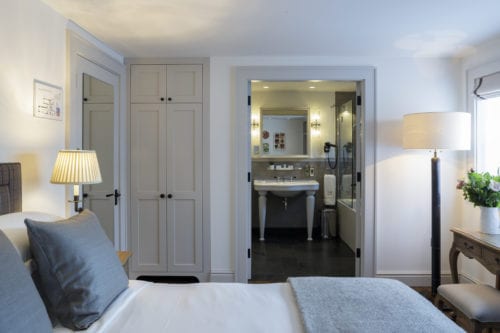 Hurley House - A cozy bedroom with a neatly made bed featuring gray pillows and a blanket. Theres a bedside lamp, a wardrobe, and an open door leading to a bathroom with a sink and mirror. Natural light filters through a window with a plant on a nearby table.