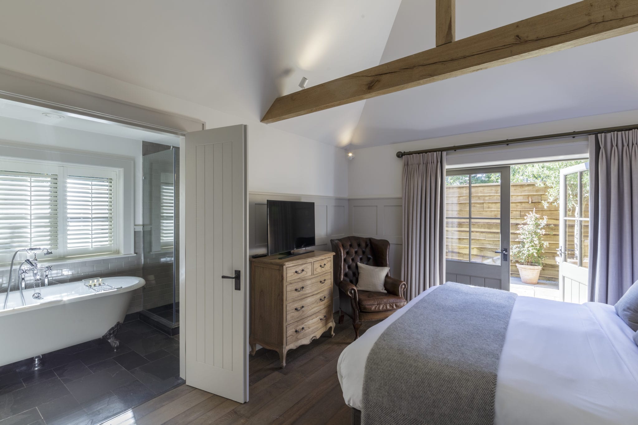 Hurley House - A cozy bedroom featuring a bed with gray and white linens, a wooden dresser with a TV, and a comfortable armchair. The room has an open bathroom with a freestanding bathtub and a door leading to a patio with wooden fencing and greenery.