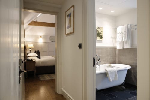 Hurley House - View of a cozy bedroom and adjoining bathroom. The bathroom features a clawfoot tub, gray tile, and neatly folded white towels. The bedroom has a wooden bed with a lamp on a nightstand. Warm lighting and wooden flooring add to the inviting atmosphere.