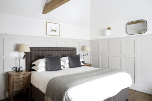 Hurley House - A cozy bedroom with a double bed featuring a checkered headboard, white and gray bedding, and two gray pillows. Two wooden nightstands with lamps flank the bed. A framed picture, an oval mirror, and a wall sconce decorate the white paneled walls.