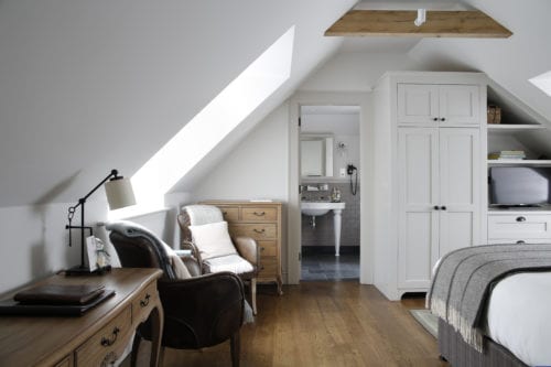 Hurley House - Cozy attic bedroom with slanted ceilings, featuring a wooden floor, white wardrobe, armchair, desk with lamp, and a view into a small bathroom. Natural light streams in through skylights, creating a serene atmosphere.