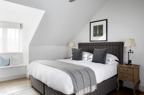 Hurley House - A cozy bedroom features a large bed with a gray upholstered headboard, adorned with white bedding and gray pillows. A soft gray throw is draped across the bed. Wooden nightstands with lamps flank the bed, and a framed picture hangs on the wall.