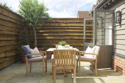 Hurley House - Outdoor patio with wooden furniture, including a table and two chairs, on stone tiles. Potted plants line the wooden fence. An open door leads inside. The scene is bright and inviting under a clear sky.