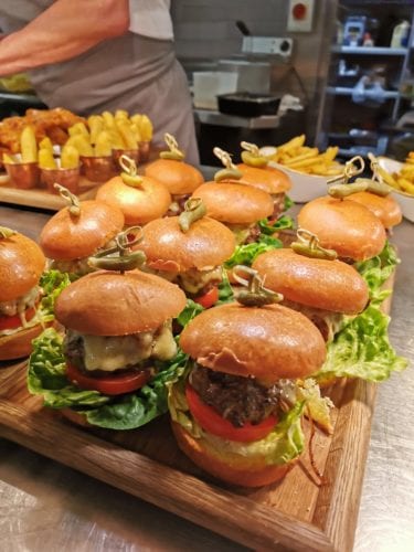 Hurley House - A wooden tray of mini burgers with lettuce, tomato, and melted cheese. Each burger is topped with a small green pepper and secured with a skewer. In the background, there are small bowls of golden fries.