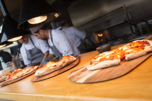 Hurley House - Pizzas on wooden boards in a restaurant kitchen with chefs working in the background under warm, hanging lights.