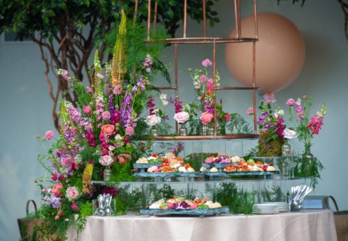 Hurley House - A beautifully arranged floral display with vibrant pink, purple, and green flowers surrounds a tiered tray of colorful, assorted appetizers on a table. In the background, theres a large peach-colored balloon and lush greenery. Plates and cutlery are on the side.