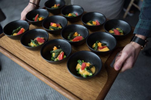 Hurley House - A person holds a wooden tray with multiple small black bowls containing a colorful dish. Each bowl appears to have greens, slices of red fruit, and an orange garnish, presented on a light gray carpeted floor.