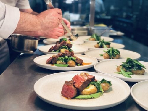 Hurley House - A chef is plating a gourmet dish of sliced steak, green vegetables, and potatoes on white plates in a professional kitchen. Other plates are arranged in a row, ready to be served.
