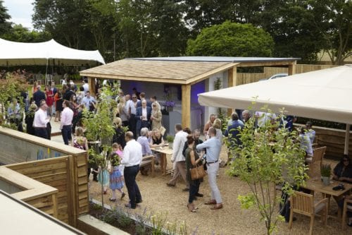 Hurley House - A lively outdoor gathering with people mingling around tables in a garden setting. Theres a wooden structure in the center, trees and tents in the background, and various individuals are engaged in conversation and socializing.