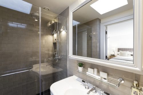 Hurley House - Modern bathroom with beige tiled walls, a glass-enclosed shower, and a white sink. A shelf above the sink holds toiletries and a small potted plant. A mirror reflects a glimpse of a bed with white linens in an adjacent room.