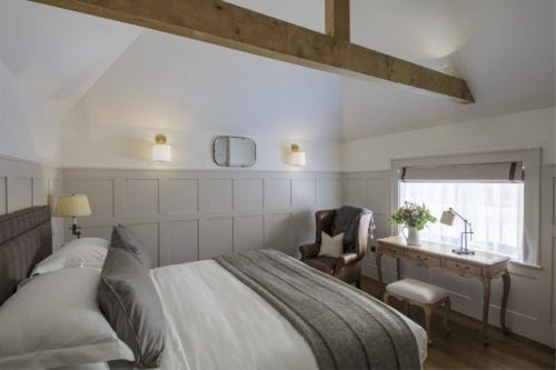 Hurley House - A cozy bedroom with a white bed, gray blanket, and wooden beams. Theres a leather armchair, desk, and stool near a window with a blind. Two wall sconces and a small mirror add lighting and decor to the paneled walls.