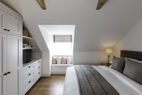 Hurley House - A cozy bedroom with white walls and wooden beams features a neatly made bed with gray accents, a window seat with cushions, a white wardrobe with shelves containing a TV and baskets, and a bedside lamp. Natural light fills the room.