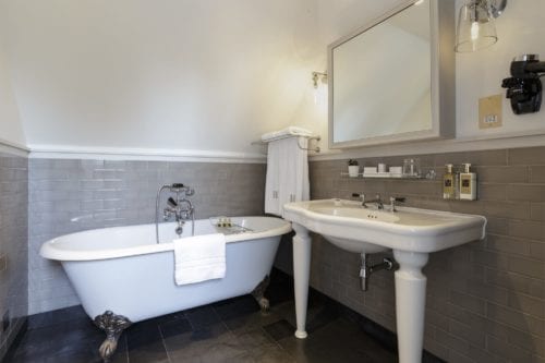 Hurley House - A bathroom with a white clawfoot bathtub and a white pedestal sink. The walls are tiled in gray, and theres a large mirror above the sink. A towel hangs on a rack, and various toiletries are placed around.