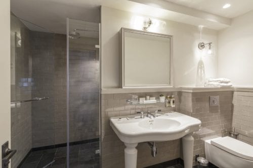Hurley House - Modern bathroom with a white sink on a pedestal, a large mirror above, and a shower with a glass door on the left. Beige tiles cover the walls, and a white toilet is on the right. Shelves hold towels and toiletries, and the lighting is bright.