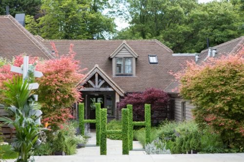 Hurley House - A charming house with a steep gabled roof is surrounded by lush gardens. The path leading to the entrance is lined with green hedges and colorful trees, set against a backdrop of dense greenery.