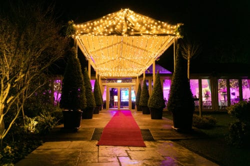 Hurley House - A nighttime scene of an elegant outdoor event space with a red carpet leading to an entrance. The path is lined with potted topiary trees, and the canopy above is adorned with sparkling string lights. The interior is illuminated with purple lighting.
