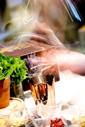 Hurley House - A blurred motion image showing a persons hand stirring a cocktail in a metallic shaker. Surrounding it are various herbs and spices, including a potted plant, adding a dynamic and vibrant atmosphere.