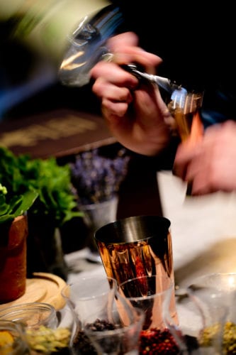Hurley House - A person vigorously shakes a cocktail shaker surrounded by various ingredients and garnishes, including herbs and spices, on a countertop. The motion creates a blur, emphasizing the dynamic action.