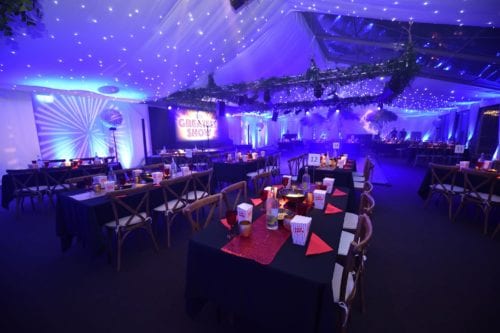Hurley House - A beautifully decorated event space illuminated in blue and purple lighting. Tables are set with snacks and drinks, while greenery and string lights adorn the ceiling. A CREATIVE SHOW neon sign is visible in the background.