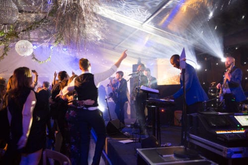 Hurley House - A lively party scene with a band performing on stage, featuring a keyboardist and guitarist in blue jackets. Attendees dance energetically, and the atmosphere is enhanced by bright lights, disco balls, and hanging decorations under a transparent tent roof.