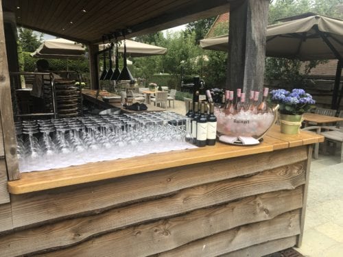Hurley House - Outdoor bar setup featuring rows of wine glasses, bottles of red wine, and a large bowl of ice holding several bottles of rosé. Wooden bar counter under a shaded pavilion, with outdoor seating and greenery in the background.